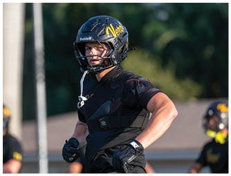 A person wearing a black football helmet and protective gear stands outdoors with a focused expression and a blurred background of trees and another person in similar gear. Descripci n generada con IA