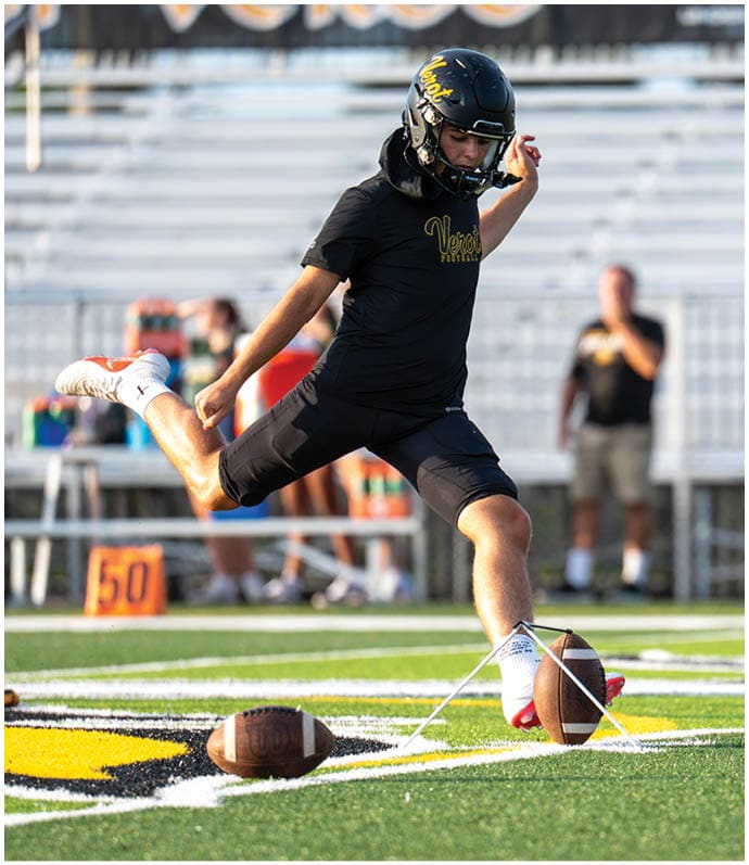 A person wearing a helmet and sportswear is kicking a football on a field while another person stands nearby watching. Descripci n generada con IA