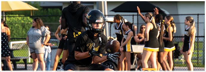 An adult in black sports gear is catching a football on a field with a group of people in cheerleading uniforms standing near a marker showing the number 20. Descripci n generada con IA