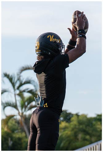 A person wearing a black football uniform and helmet is jumping to catch a football outdoors near bleachers and palm trees. Descripci n generada con IA