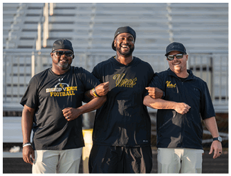 Three adults wearing casual sportswear and caps stand linked arm in arm smiling on a sports field with bleachers in the background. Descripci n generada con IA