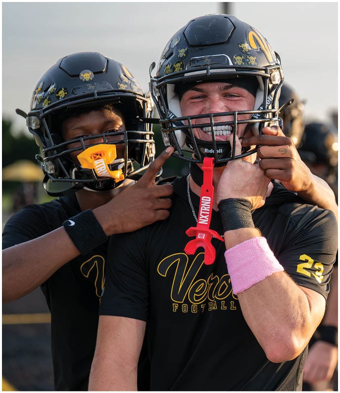 Two people wearing football helmets and black jerseys are playfully interacting on a football field during sunset. Descripci n generada con IA
