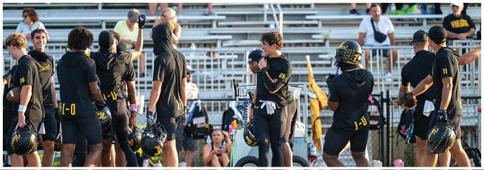 People in sports uniforms stand and sit on a field near bleachers, some holding helmets and others appearing to listen or prepare for an activity. Descripci n generada con IA