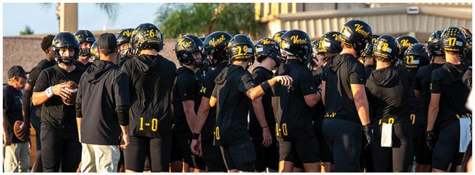 A group of people wearing black athletic clothing and helmets are gathered closely together on a grassy field, appearing to be part of a sports team. Descripci n generada con IA