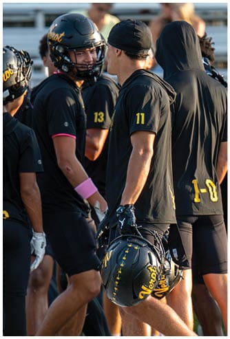 People wearing black sports uniforms with yellow numbers are standing and holding football helmets on a field. Descripci n generada con IA