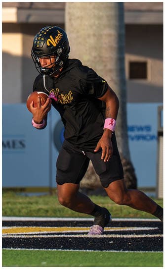 A person wearing a black sports uniform and helmet is running on a football field near a trash can and a building in the background. Descripci n generada con IA