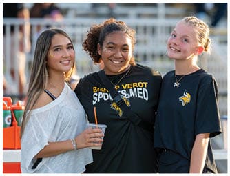 Three people smiling and standing close together outdoors with one holding a drink and wearing casual clothing including sports-themed shirts. Descripci n generada con IA
