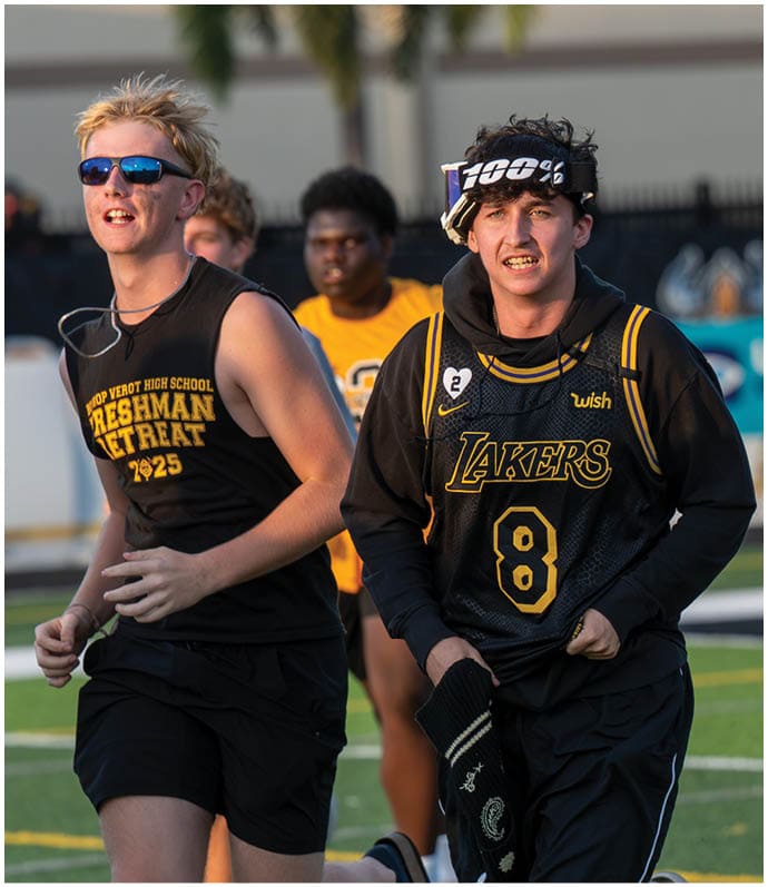 A person wearing sunglasses and a sleeveless black shirt with “Freshman Retreat 2025" printed on it is smiling outdoors with other people in the background. Descripci n generada con IA