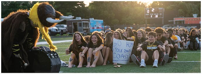 A group of people sitting on a sports field with one person holding a sign that says “GO HOME Your own COAST" while a mascot in a costume interacts with them. Descripci n generada con IA