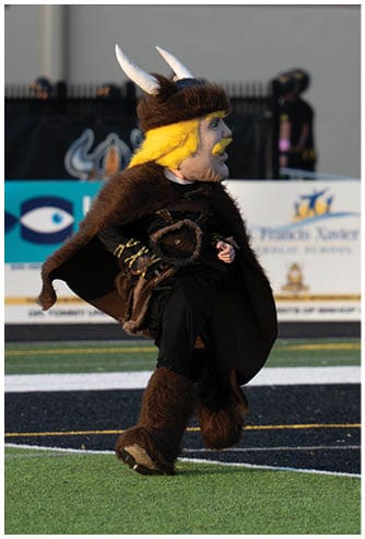 A person in black clothing is running on a sports field while another person nearby is holding a helmet and walking. Descripci n generada con IA