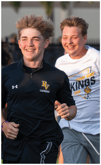 A person with tousled hair wearing a black sports shirt with a lacrosse logo is smiling and running outdoors with others in the background. Descripci n generada con IA