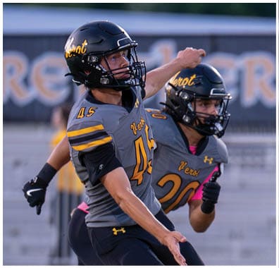 Two people in football uniforms and helmets are actively engaged in a game on a field with bleachers and a banner in the background. Descripci n generada con IA