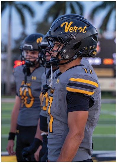 Two people wearing gray and yellow football uniforms and helmets stand on a field with palm trees in the background. Descripci n generada con IA