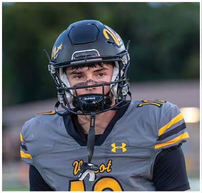A person wearing a black football helmet and gray football jersey with yellow accents stands outdoors with a focused expression. Descripci n generada con IA