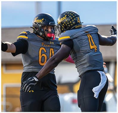 Two people in football uniforms celebrating on a field with one extending arms toward the other. Descripci n generada con IA