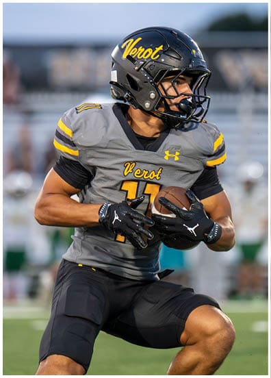 A person wearing a gray and black football uniform with the word “Verot" on the helmet and jersey is holding a football and appears to be running on a field during a game. Descripci n generada con IA