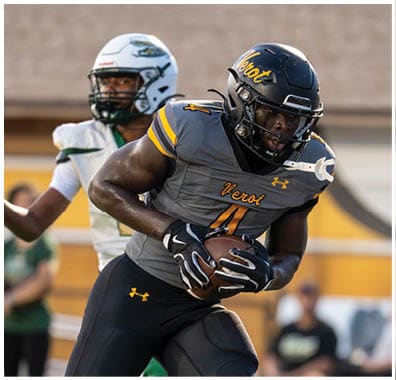 A person in a dark football uniform with “Verot" on the jersey is running with a football while another person in a white football uniform is reaching out behind them. Descripci n generada con IA