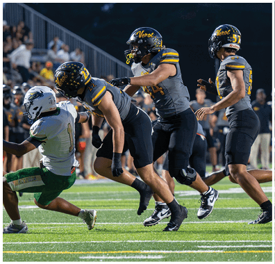 Three football players in gray and black uniforms tackle a player in a white and green uniform on a field during a night game. Descripci n generada con IA