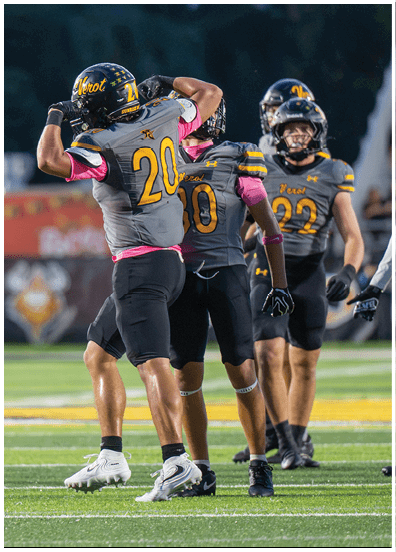 Three people in football uniforms are on a field with one person flexing their arms while the others stand nearby. Descripci n generada con IA