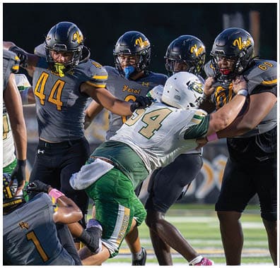 People in football uniforms are engaged in a play with one person in a white and green uniform pushing against several people in dark gray and yellow uniforms. Descripci n generada con IA