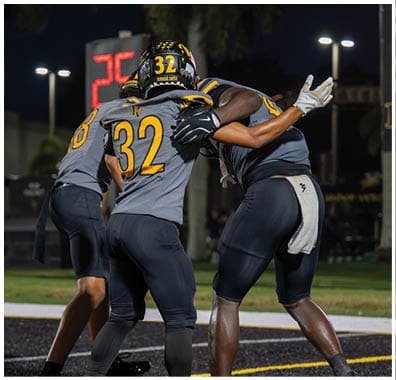 Three people in football uniforms are celebrating on a field at night with one person hugging another. Descripci n generada con IA