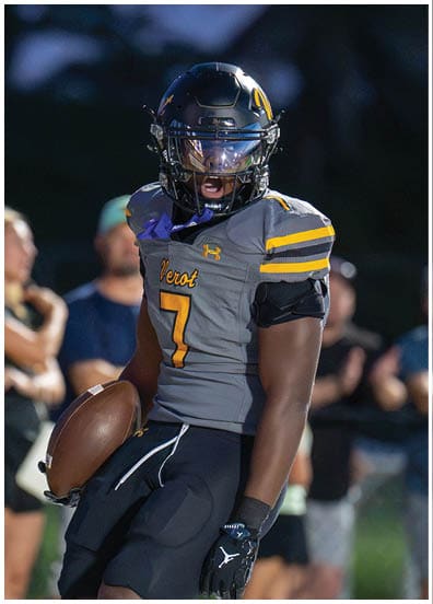 A person wearing a football uniform and helmet is holding a football and appears to be shouting or celebrating on a field with blurred spectators in the background. Descripci n generada con IA