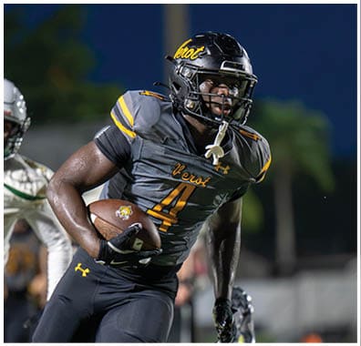 An adult wearing a black and gray football uniform with the word “Verot" on the chest is running while holding a football during a nighttime game. Descripci n generada con IA