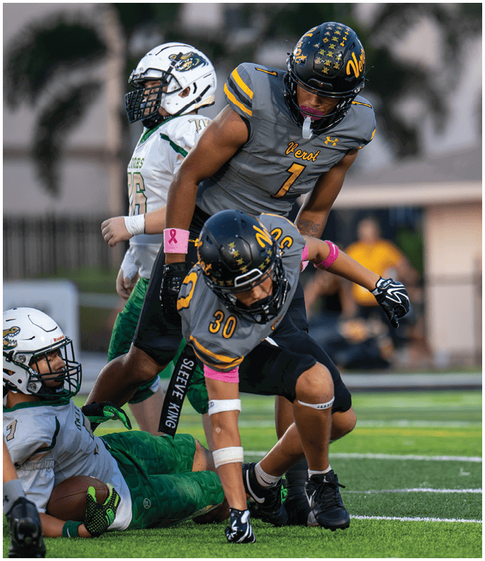Two people in gray and black football uniforms with pink wristbands are engaged in a play on a football field while a person in a white and green uniform and a referee stand nearby. Descripci n generada con IA