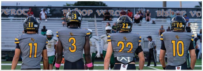 Four people wearing football uniforms with helmets stand side by side on a field facing away from the camera toward empty bleachers. Descripci n generada con IA