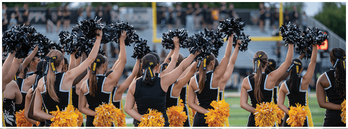 A group of people in matching black and yellow cheerleading uniforms are standing in a line on a sports field, holding black pom-poms raised above their heads and yellow pom-poms at their sides. Descripci n generada con IA