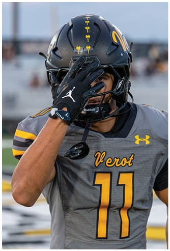 A person wearing a football helmet and gray jersey with the number 11 and the word Verot is adjusting their helmet with a gloved hand. Descripci n generada con IA