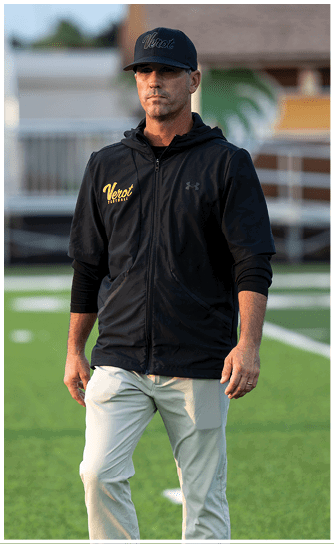 A person wearing a black zip-up hoodie, light-colored pants, and white athletic shoes is standing on a green sports field. Descripci n generada con IA