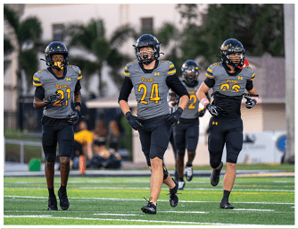 Four people wearing football uniforms with helmets are running on a green field during a game or practice. Descripci n generada con IA