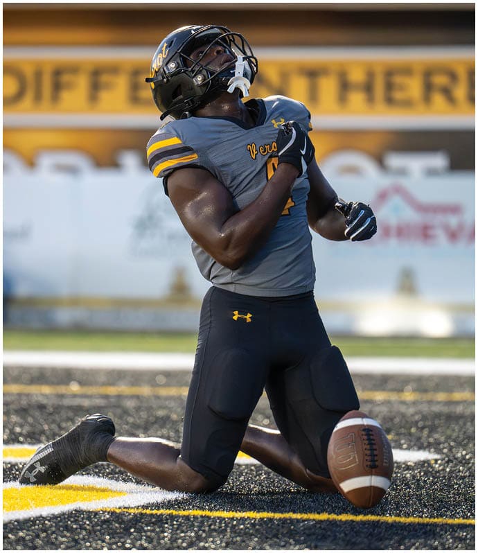 A person wearing a football uniform kneels on the field with their head tilted back and arms raised near their chest next to a football. Descripci n generada con IA