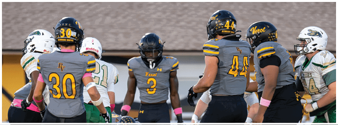 Five people in football uniforms are standing on a field facing each other, with some wearing gray and yellow jerseys and others in white and green jerseys. Descripci n generada con IA