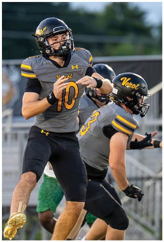 Two people in football uniforms and helmets are actively engaged in a game on a field with a blurred background of trees and structures. Descripci n generada con IA