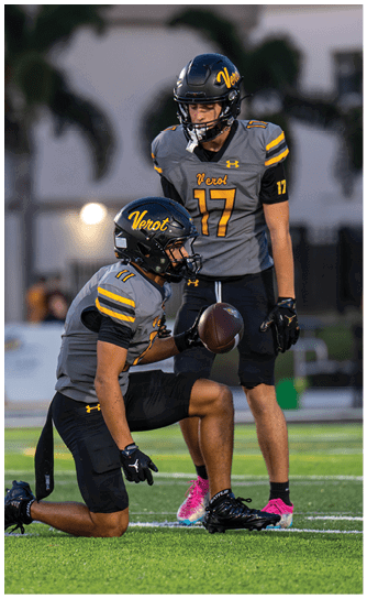 Two people wearing gray and black football uniforms with helmets are on a football field, with one kneeling and holding a football while the other stands behind. Descripci n generada con IA