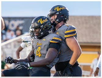 Two people wearing football helmets and gray jerseys with yellow lettering celebrate on a field during a game. Descripci n generada con IA