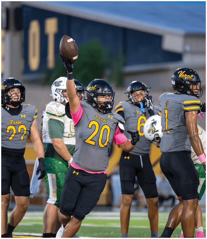 A person wearing a football helmet and gray jersey with the number 20 is raising a football in the air while another person in a similar uniform stands nearby. Descripci n generada con IA