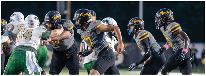 Several people in football uniforms are engaged in a play on a field, with some players in dark uniforms blocking or running against others in white and green uniforms. Descripci n generada con IA