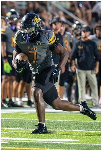 A person in a football uniform is running with the ball on a field while another person in a different uniform reaches out toward them, with a crowd of people watching in the background. Descripci n generada con IA