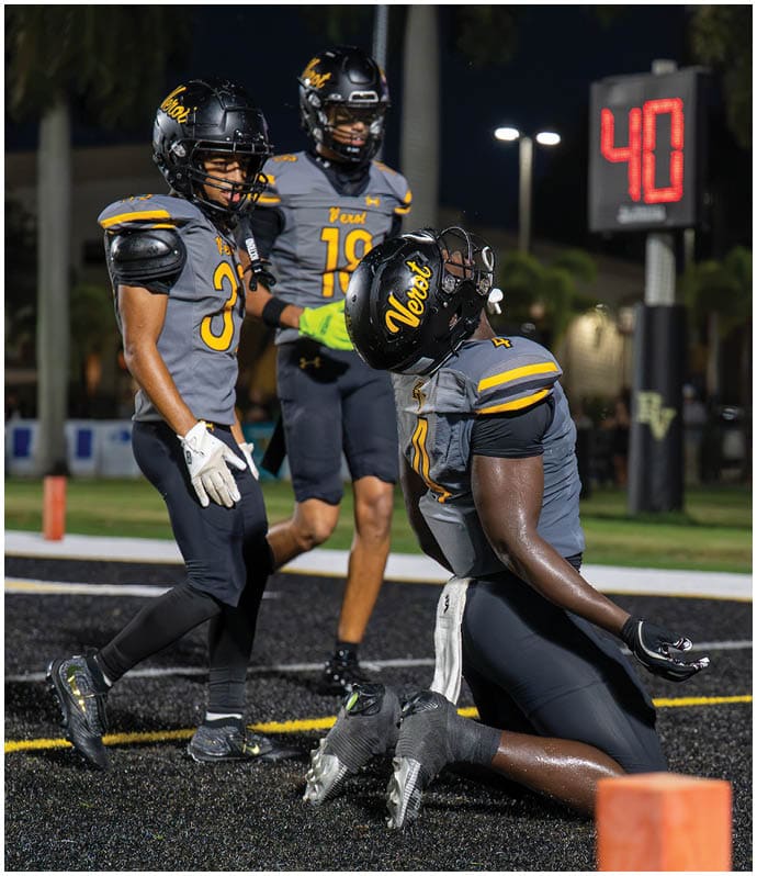 A person in a football uniform kneels and leans back in celebration near the end zone while two other players stand nearby on a nighttime field. Descripci n generada con IA