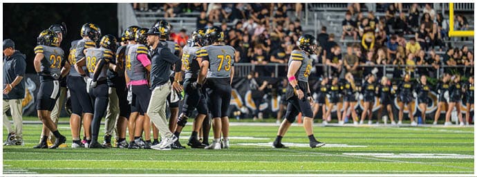 A group of football players and coaches in black and yellow uniforms stand on the field with a crowd in the background during a game. Descripci n generada con IA