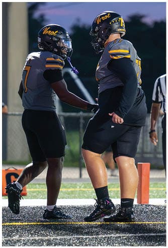 A person in a football uniform and helmet stands on a field near an orange pylon while a referee in a striped shirt watches in the background. Descripci n generada con IA