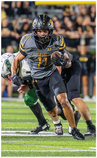 A person in a black and yellow football uniform is running with the ball while another person in a white helmet and green pants attempts to tackle them on a football field with a crowd in the background. Descripci n generada con IA