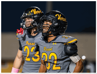 Two people wearing gray and yellow football uniforms and helmets are on a field, with one person in the foreground looking upward and the other in the background making a gesture with their hand. Descripci n generada con IA