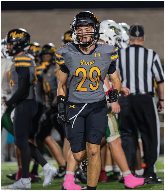 A person wearing a black helmet and gray football jersey with the number 29 stands on a field with two other people in white helmets and a referee in a black and white striped shirt in the background. Descripci n generada con IA