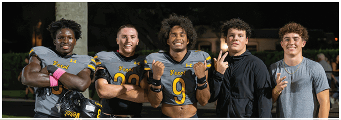 Five people stand close together at night with three wearing football uniforms and smiling while making hand gestures. Descripci n generada con IA
