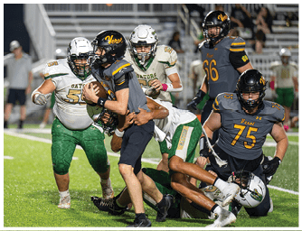 A group of people in football uniforms are engaged in a tackle on a grassy field during a game. Descripci n generada con IA