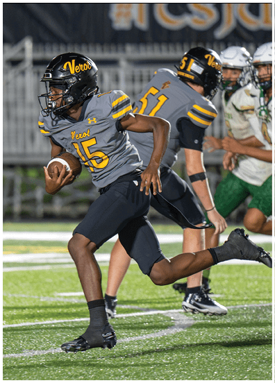 A person wearing a gray and black football uniform with the number 15 is running on a field while holding a football, with other players visible in the background. Descripci n generada con IA